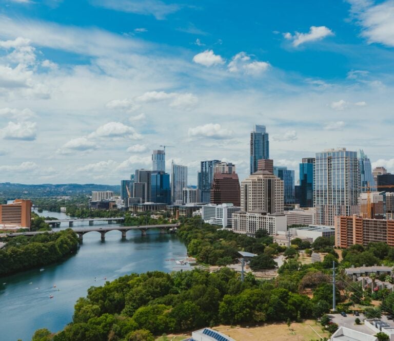 An aerial view on a sunny and white puff cloud day of the Austin, TX city skyline with Lady Bird Lake running beside it