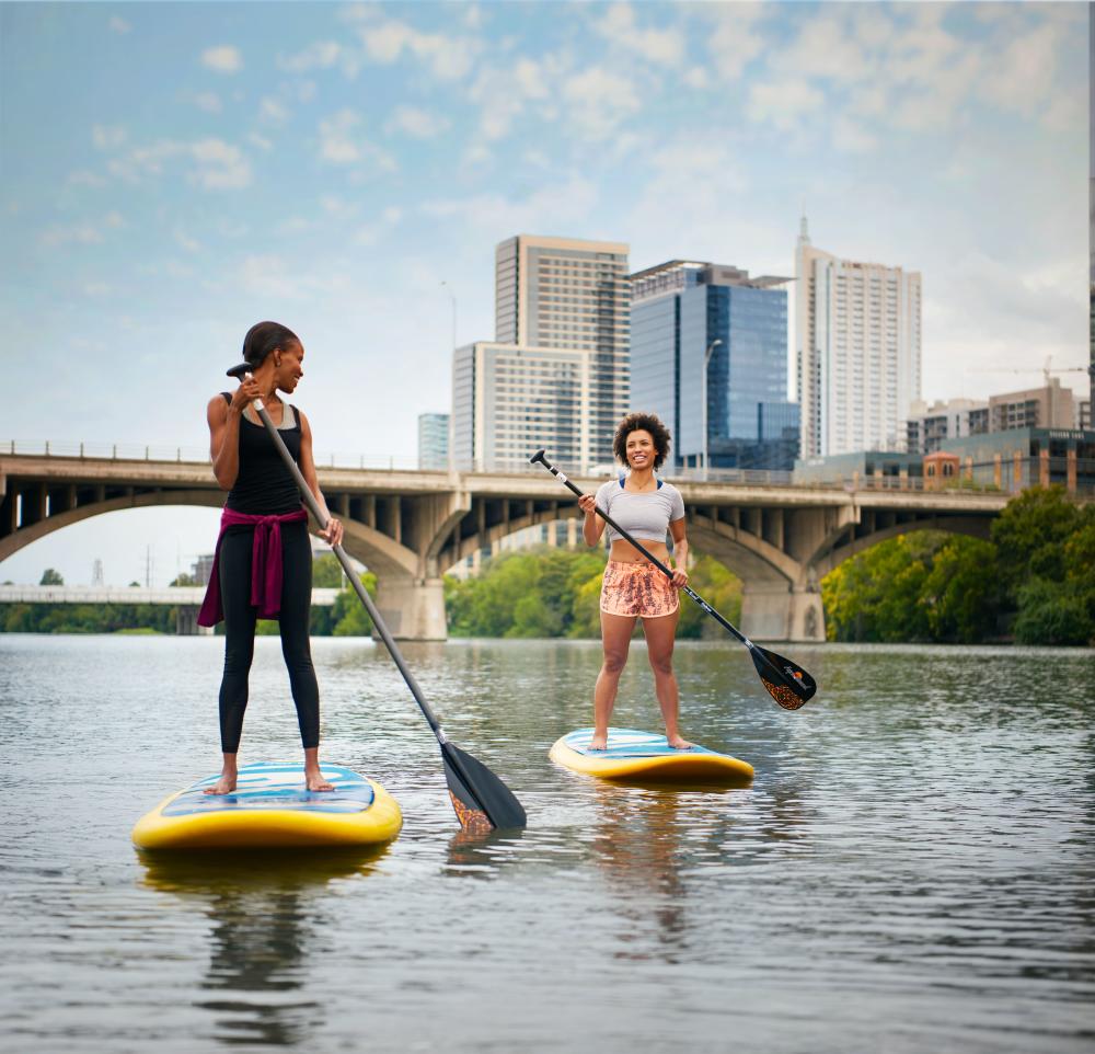 Too young women enjoy paddleboarding on a beautiful day on Lade Bird Lake with the Austin, TX city skyline in the background