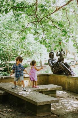 Children smile while playing on the concrete seating in the wooded Umlauf Scupture Garden grounds