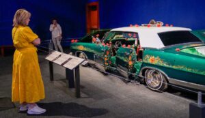 1967 Ford LTD lowrider car. Painted with bright green paint with rose details and a white roof. Woman in a long bright yellow dress standing beside it looking at the display.