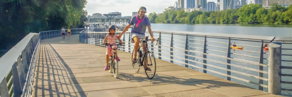 A man on a bike and his daughter enjoy a walk and bike ride on the Butler Hike-and-Bike Trail at Lady Bird Lake, Austin