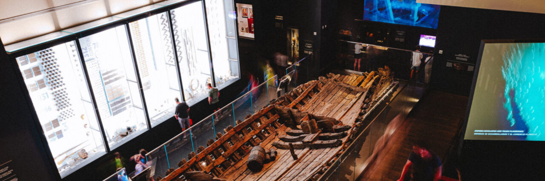 Modern museum interior featuring a large preserved wooden shipwreck exhibit surrounded by glass railings, with visitors walking through interactive digital displays and illuminated informational panels in a dimly lit gallery.