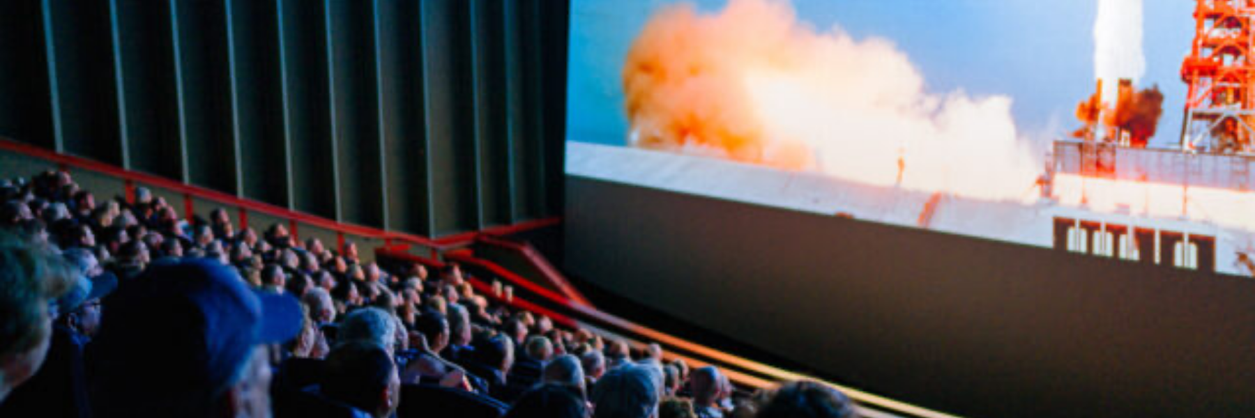 Crowded movie theater audience seated in rows watching a large cinema screen displaying a rocket launch explosion scene, with dramatic lighting reflecting off viewers in a dark auditorium.