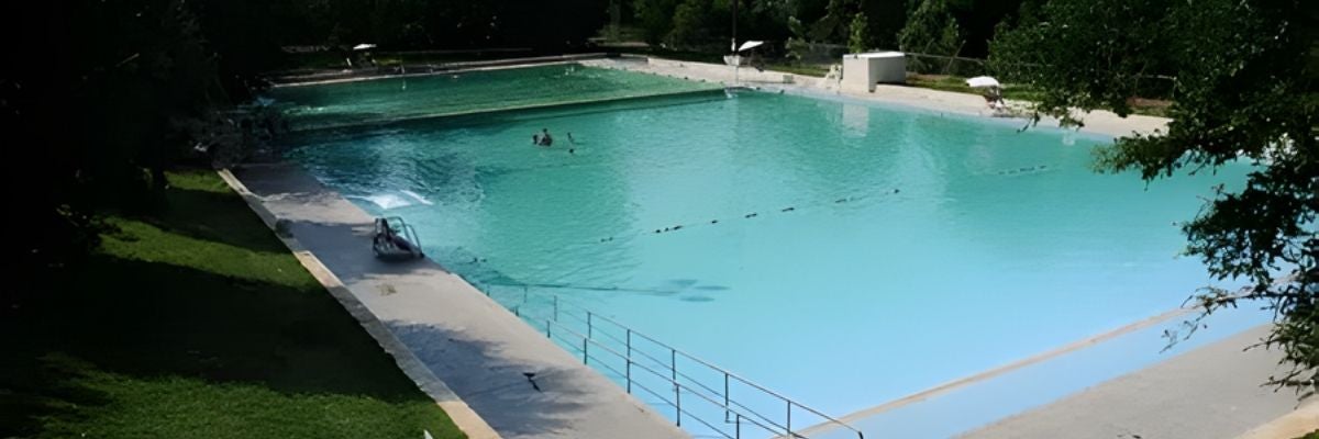 A large, tranquil outdoor pool with clear blue water, surrounded by trees and grassy areas. A few people swim, evoking a calm, summery vibe.