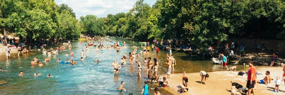 A lively summer scene at a river with many people swimming and relaxing on the banks. The area is lush with trees, creating a vibrant, joyful atmosphere.