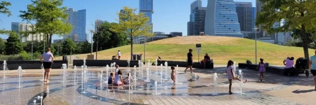 Children play in a splash pad with water jets in the Liz Carpenter Fountain at Butler Park in Austin, TX. The Austin city skyline towers in the background with trees and the park grounds.