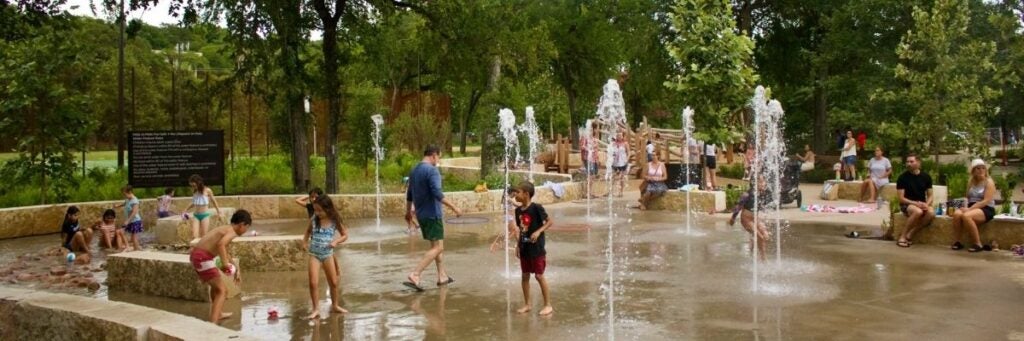 Children and adults play in the Pease Park splash pad with water jets, surrounded by trees and benches in Austin, TX.