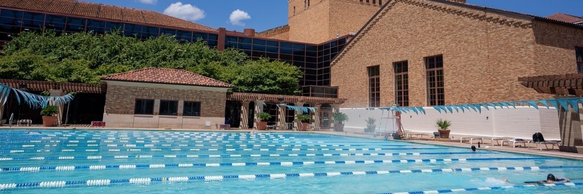 Outdoor swimming pool with blue lanes, surrounded by brick buildings and lush greenery under a clear blue sky. A calm, leisurely atmosphere.