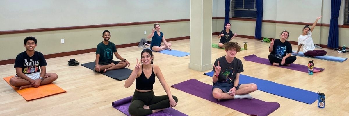 Three people meditate on yoga mats in a serene park. They sit cross-legged with eyes closed, surrounded by autumn leaves and trees, conveying tranquility.