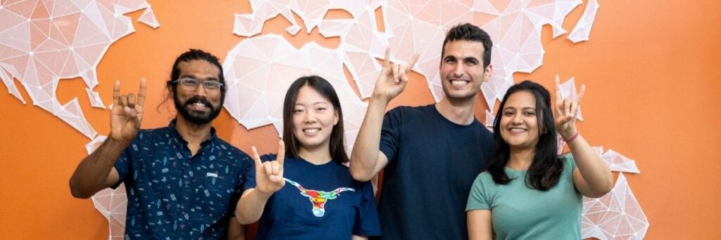 A diverse group of four people smiling and making the Longhorn symbol in front of a geometric world map on an a UT official orange background.