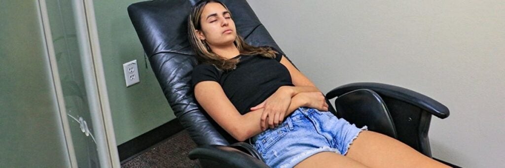 A young women reclining on a black leather chair with eyes closed, wearing a black t-shirt and denim shorts.
