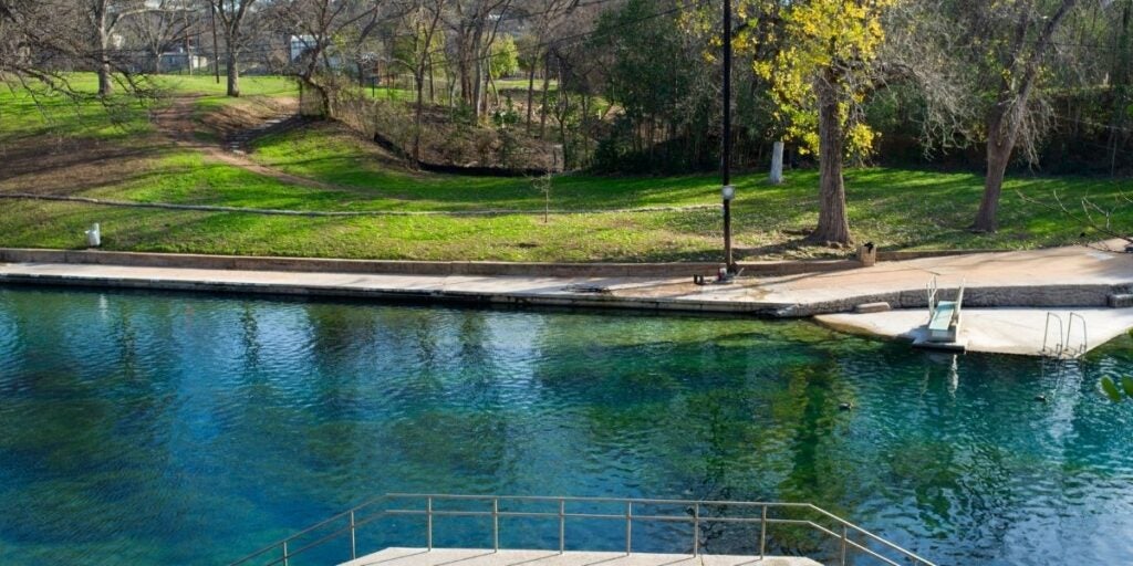 Deep Eddie, a natural outdoor pool with clear blue-green water, bordered by a concrete walkway, adjacent to a grassy park with trees.