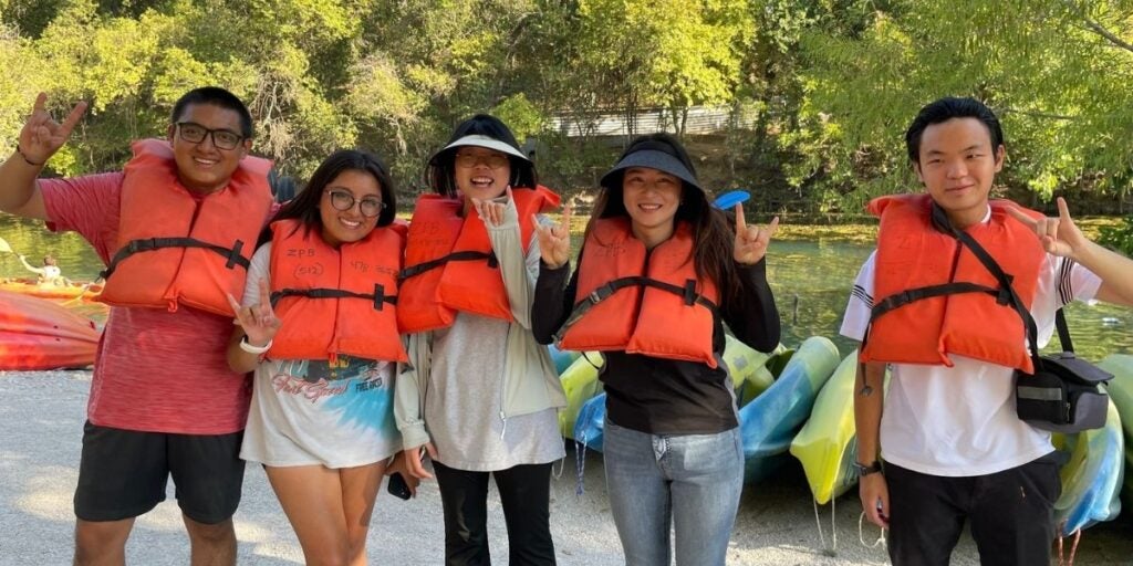 Five ELC students wearing orange life jackets standing in front of kayaks and trees, smiling.