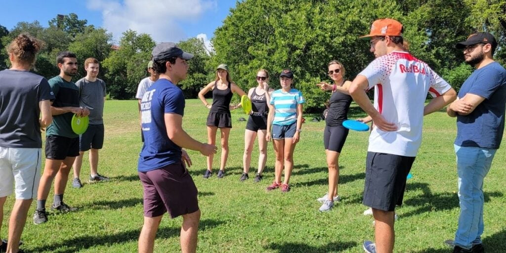 A group of students in sportswear stand in Zilker Parkl with frisbees, surrounded by trees.