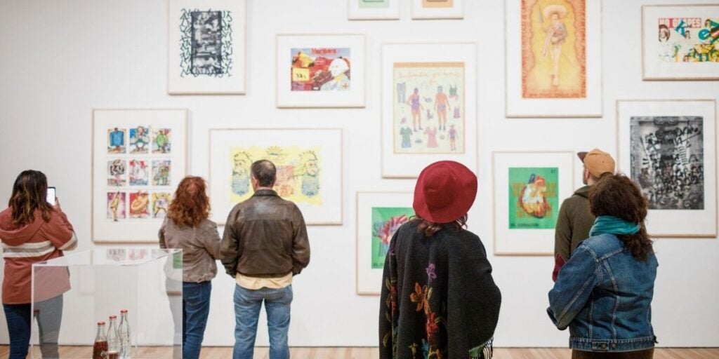 The image shows a gallery setting with several people looking at an art exhibit on a white wall.