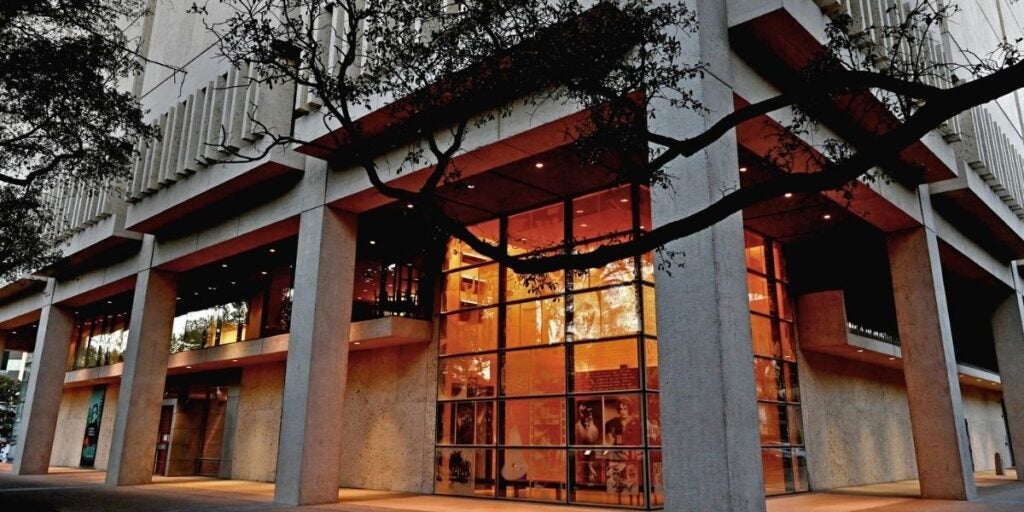 The image depicts a modern architectural building with a striking facade. It features large concrete columns supporting a flat roof, under which are large glass panels glowing with warm, orange lighting.