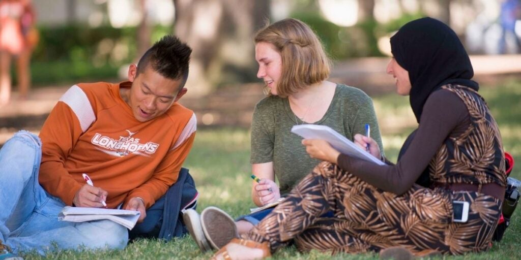 The image shows three students sitting on grass in an outdoor setting, engaging in a group study session.
