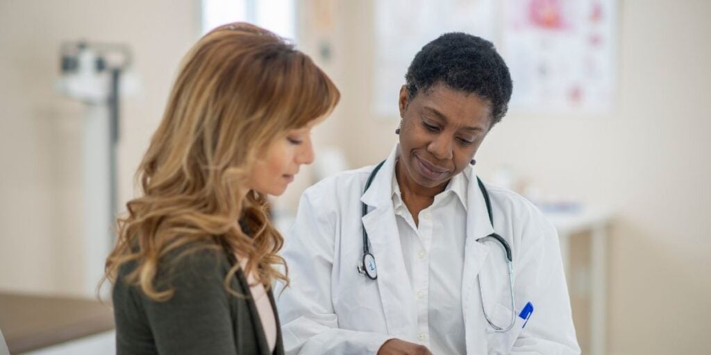 A doctor in a white coat, holding a stethoscope, consults with a patient. They're in a medical office, engaged in a serious yet caring discussion.