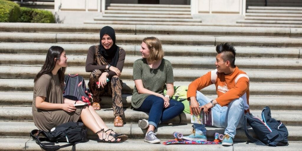 Four students sit on outdoor steps, chatting happily. They differ in attire and appearance, with backpacks and a skateboard nearby, conveying a relaxed atmosphere.