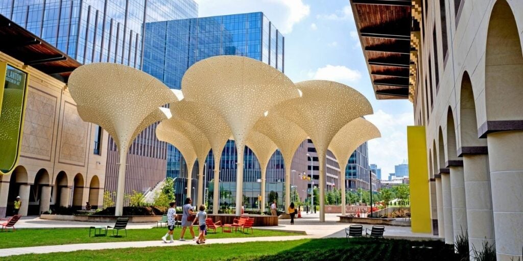UT Austin outdoor plaza with large white sculptural canopy structures, one of the many Landmarks campus art installations, surrounded by modern buildings with visitors and students passing underneath them.