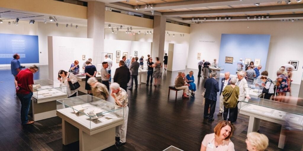 Visitors observe art and writings on display at the gallery in the UT Austin Harry Ransom Center. The gallery has a spacious interior with polished dark wooden floors and cream-colored walls. Several display cases with glass tops are positioned throughout the room.