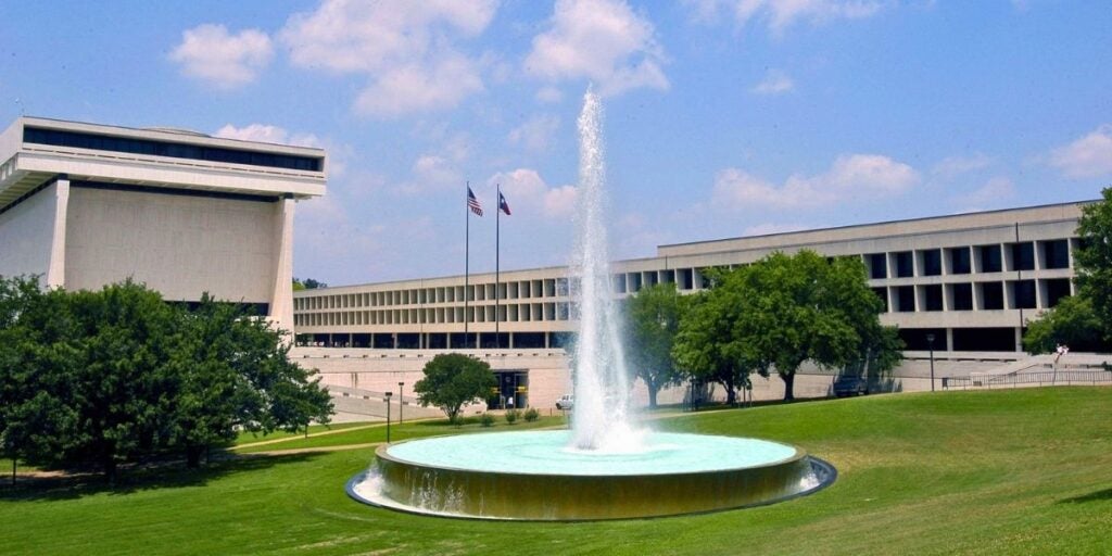 The LBJ Presidental Libary at UT Austin is seen with the circular LBJ fountain in the green concouse in front blasting a tall jet of water against the blue sky above on a sunny day on the UT campus.
