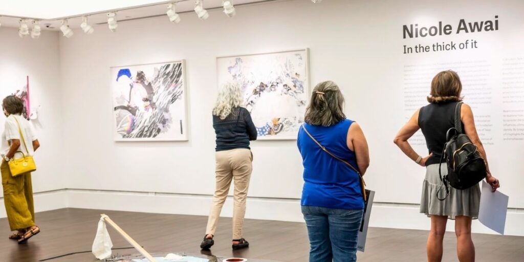 Visitors observing abstract paintings in an Art Galleries at Black Studies Exhibiton with text "Nicole Awai, In the thick of it" on the wall.