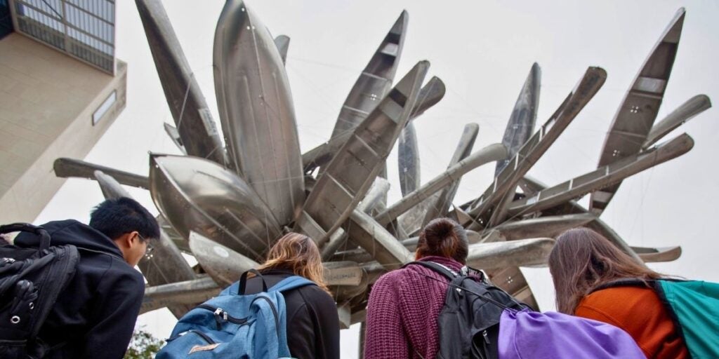 The image shows the perspective from behind a group of students, observing one of the large Landmarks Public Art sculptures on the UT Austin campus. The massive sculpture comprises numerous aluminum boats arranged in a chaotic radial pattern, pointing outward and upward like petals of a flower.