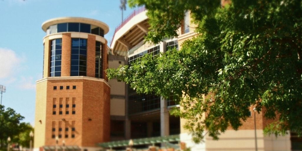 The UT Austin orange brick and glass cylindrical Stark Center is shown with an oak tree in the foreground. The Texas memorial stadium sits beside it.
