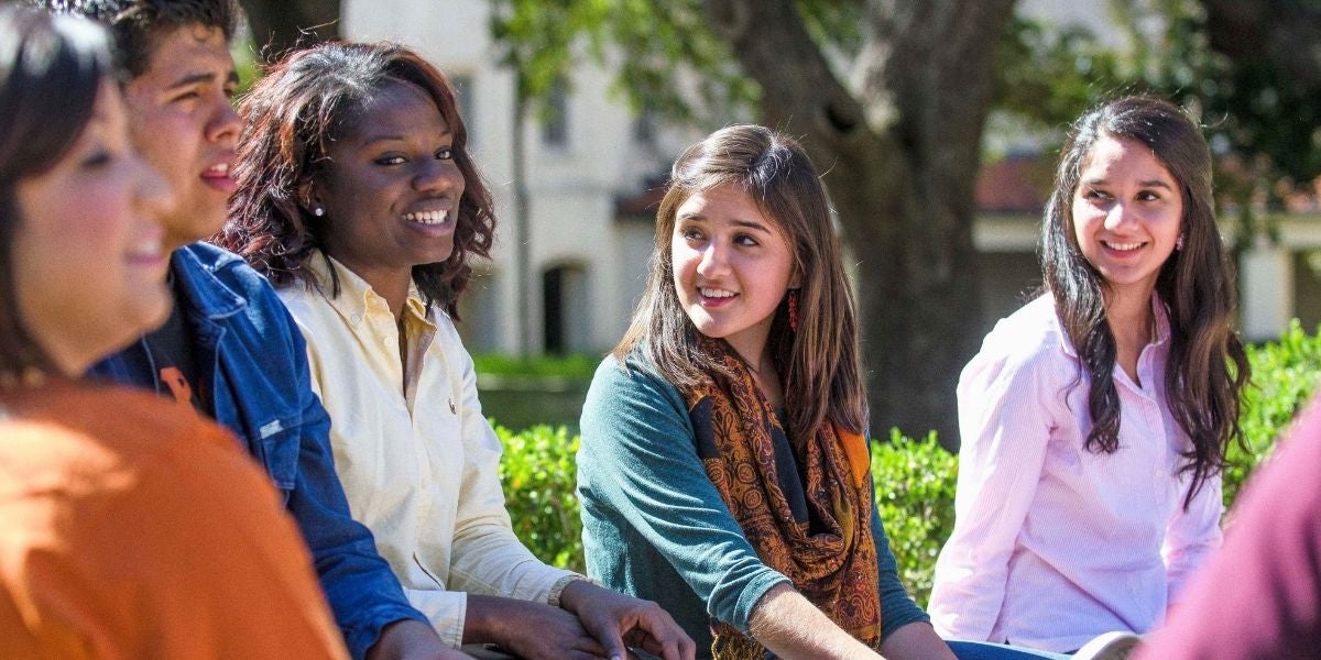 A group of five students sitting on a bench outdoors, smiling and engaging in conversation. Sunlight filters through the trees, creating a warm, inviting atmosphere.