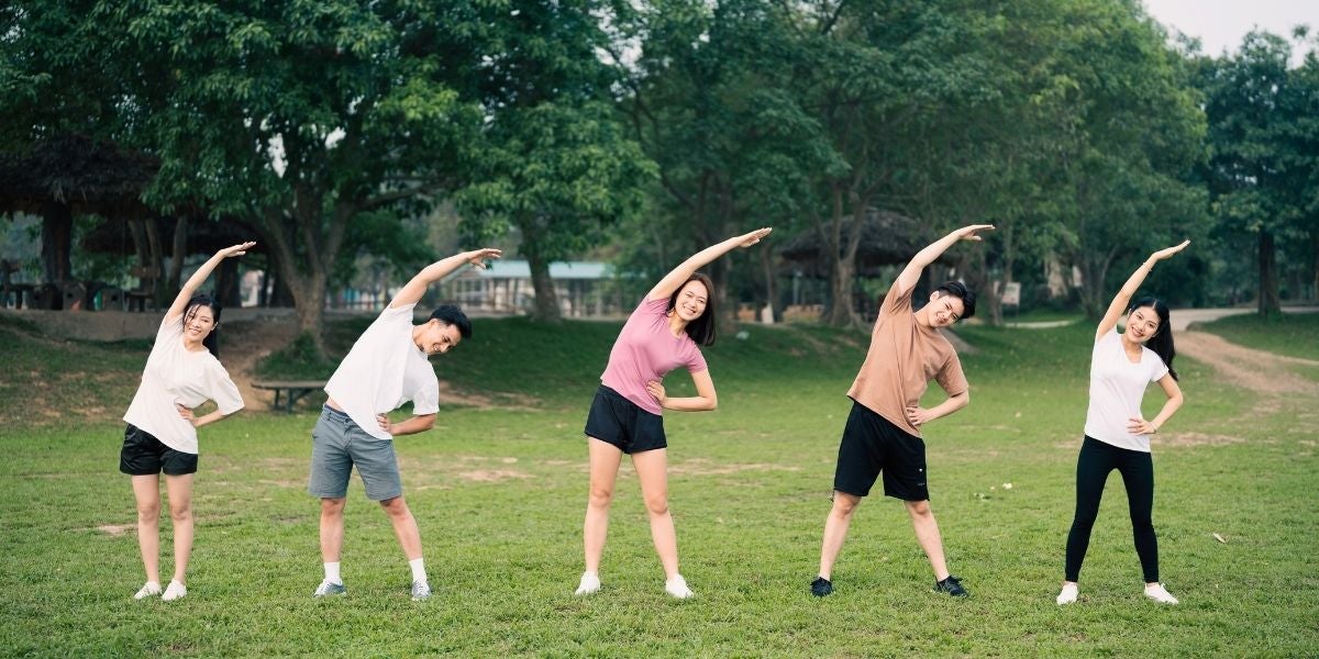 Five people stretch side by side on a grassy field, each bending to their right with one arm overhead. The scene is relaxed and sunny, with lush trees in the background.