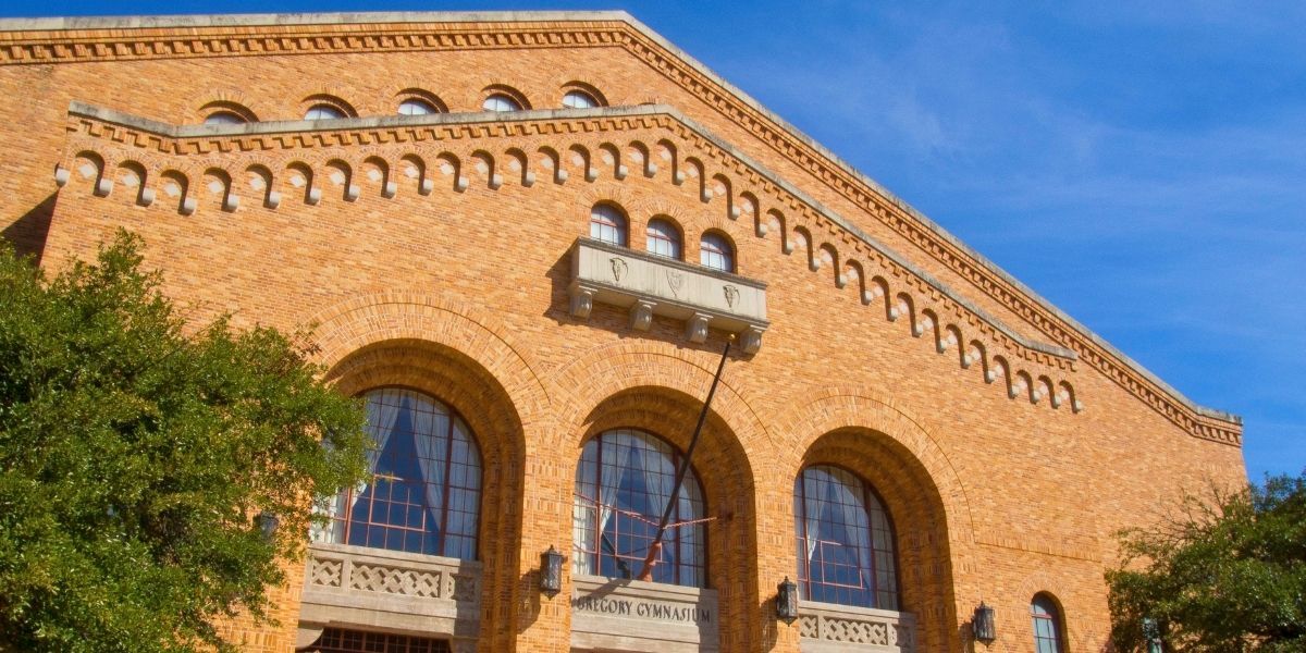 Historic brick building facade with arched windows and decorative brickwork under a clear blue sky. Trees partially frame the scene, adding greenery.