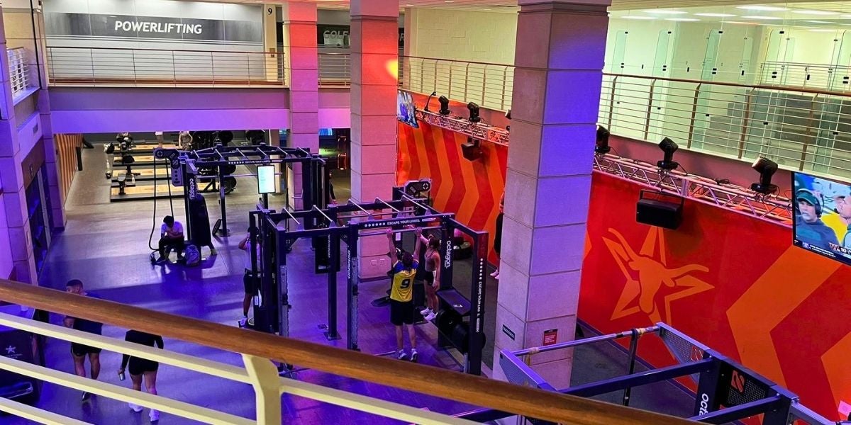 Gym interior with vibrant lighting. Powerlifting area seen from above; people exercise on equipment. Bright red-orange walls, star graphics, and TV screen.