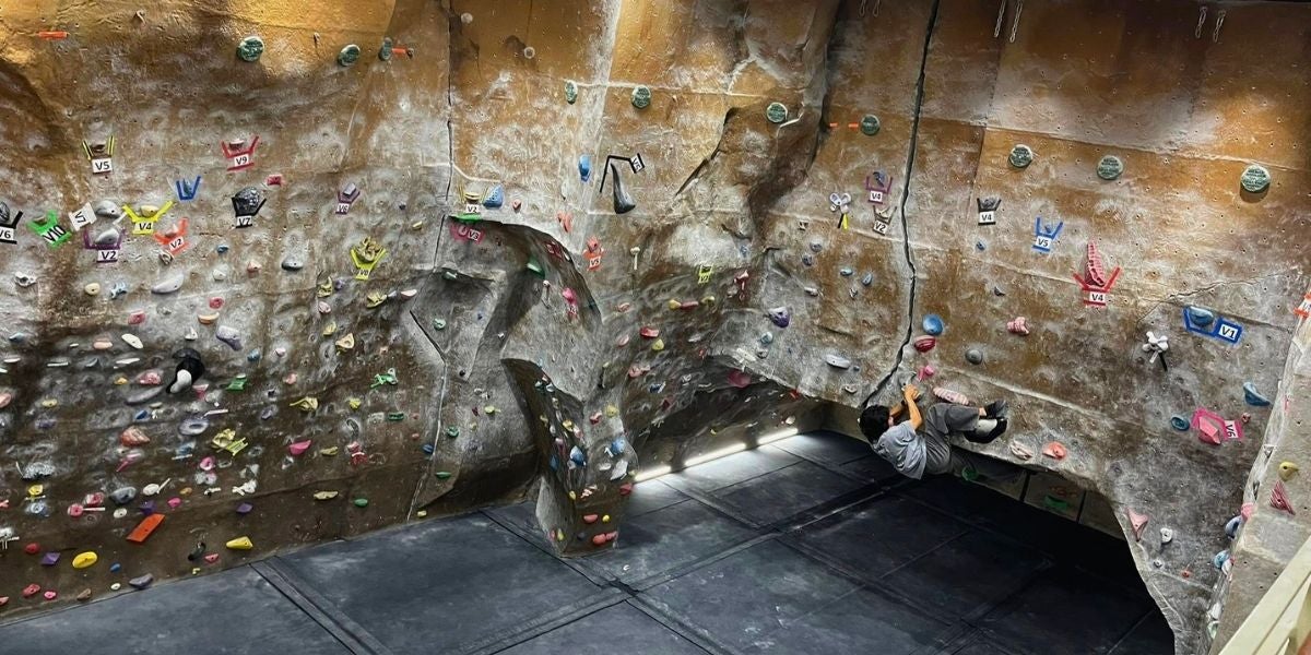 A climber navigates a textured indoor rock climbing wall with colorful holds. The scene is dynamic, conveying focus and determination.