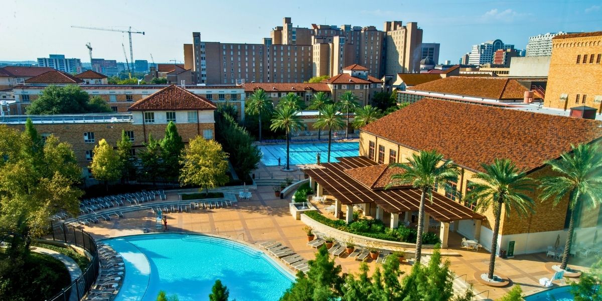 Aerial view of a sunny outdoor pool area with clear blue water, surrounded by lounge chairs, trees, and terracotta-roofed buildings. Urban skyline in the background.