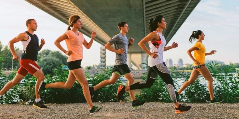 Five people jogging under a bridge on a gravel path, surrounded by greenery. They appear focused and energetic, conveying a sense of fitness and determination.