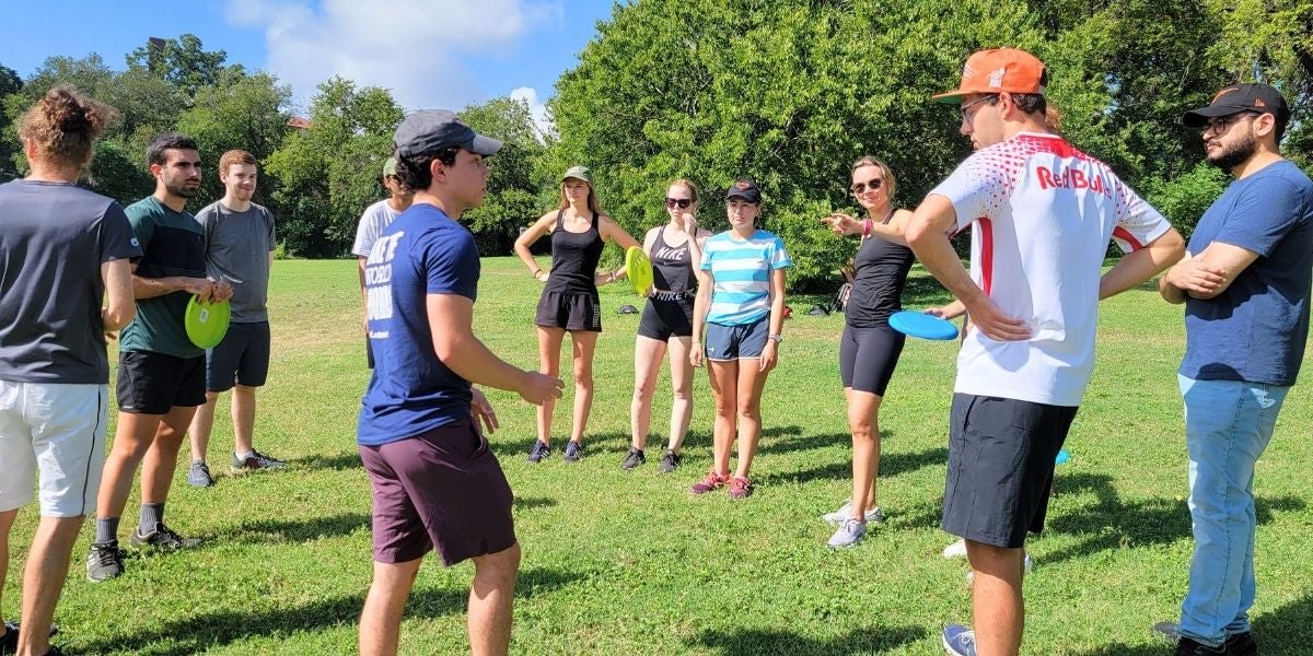 A group of people stands in a sunny park, holding frisbees. They appear engaged and attentive, possibly during a team meeting or a frisbee game briefing.