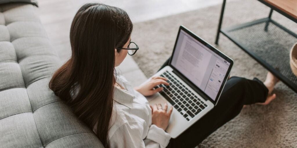 A woman sits on the floor beside a gray sofa, working on a laptop. She has long hair and wears glasses and a white shirt, conveying a focused mood.