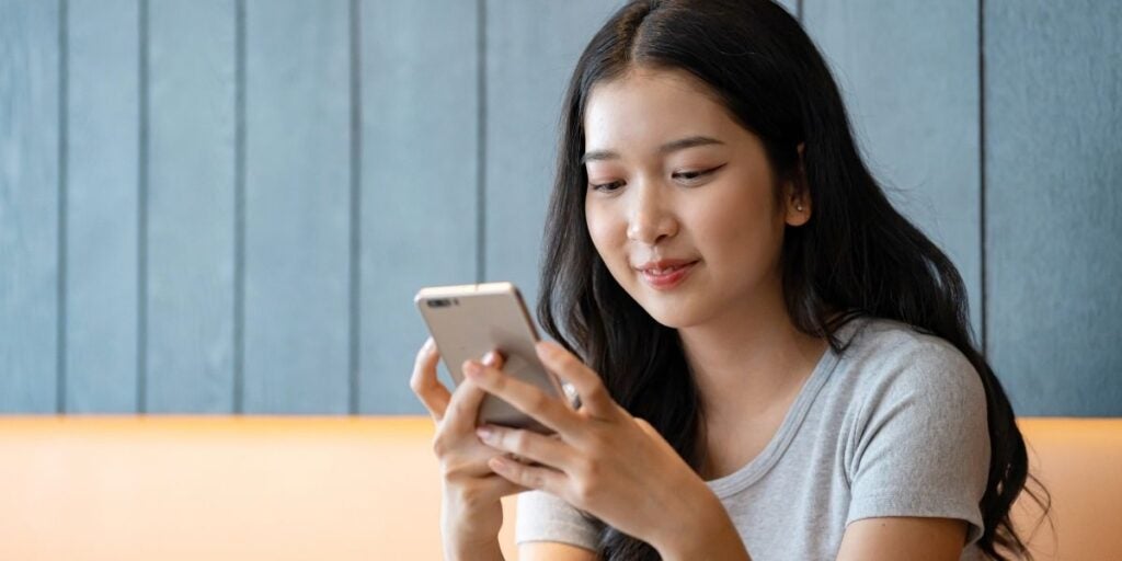 Young woman with long hair, smiling as she looks at her smartphone. She is seated indoors against a dark wood panel wall, wearing a light gray shirt.