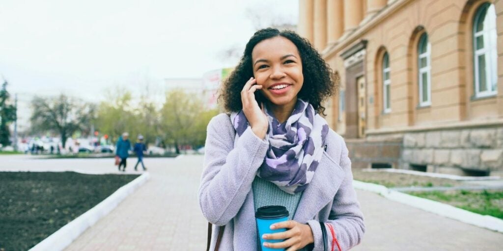 A smiling woman in a lavender coat and scarf talks on her phone while holding a blue cup. She stands on a city sidewalk near a brown building.