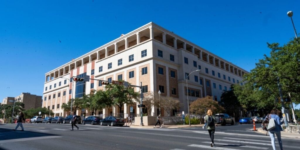 Street view of a large, multi-story beige building with a clear blue sky. People cross at a crosswalk, while cars wait at a traffic light. Trees line the sidewalk.