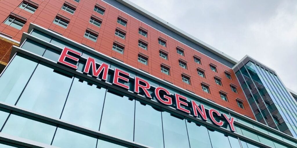 Red brick hospital building with large "EMERGENCY" sign in bold red letters. Modern glass facade, conveying urgency and healthcare readiness.
