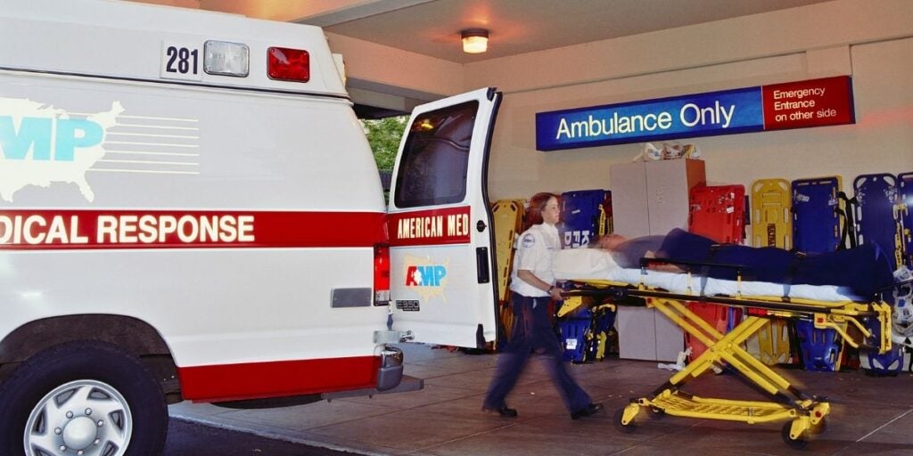 Paramedic pushes a stretcher with a patient into a brightly lit hospital emergency entrance. An open ambulance is parked beside a sign reading "Ambulance Only."