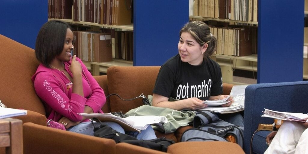 Two young women talking in a library, surrounded by books and file boxes.
