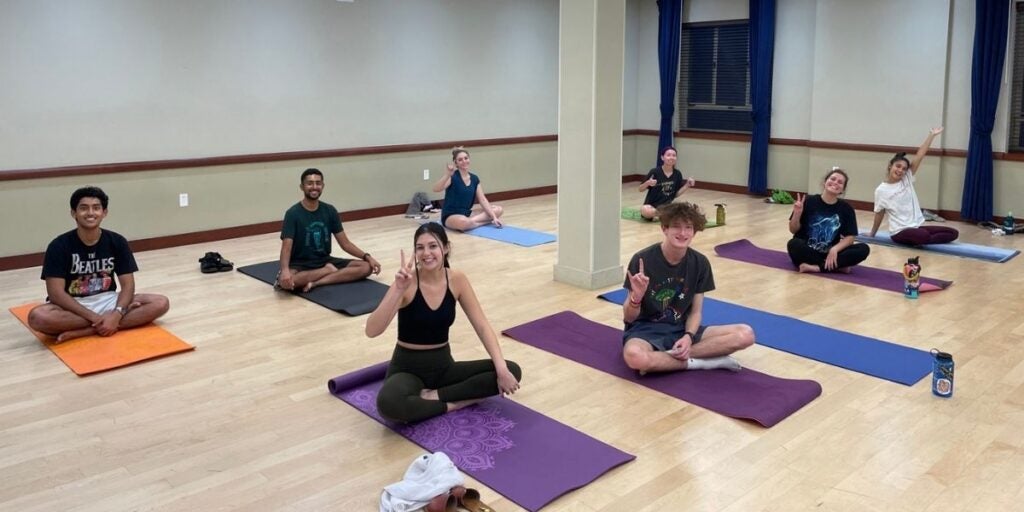 A group of eight people seated on yoga mats in a spacious room with wooden floors and beige walls, smiling and making hand gestures.
