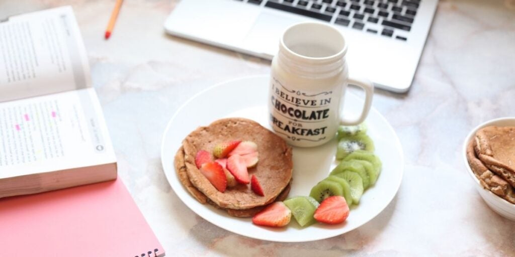Plate with pancakes, strawberries, kiwi, and a cup with text; laptop and books in the background.