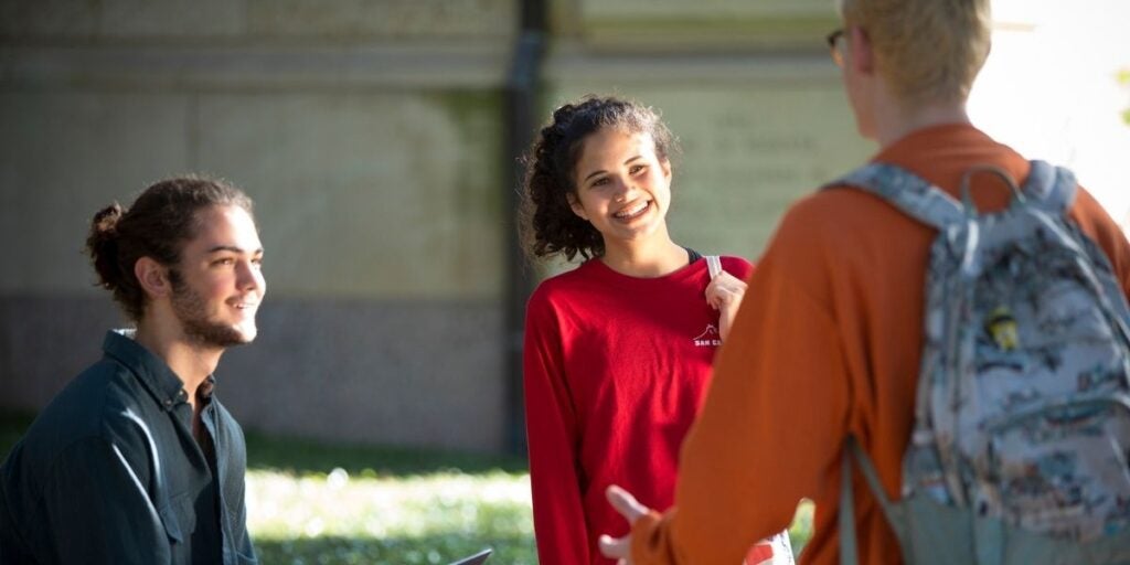 Three students talking outside on a sunny day, with one seated and two standing.