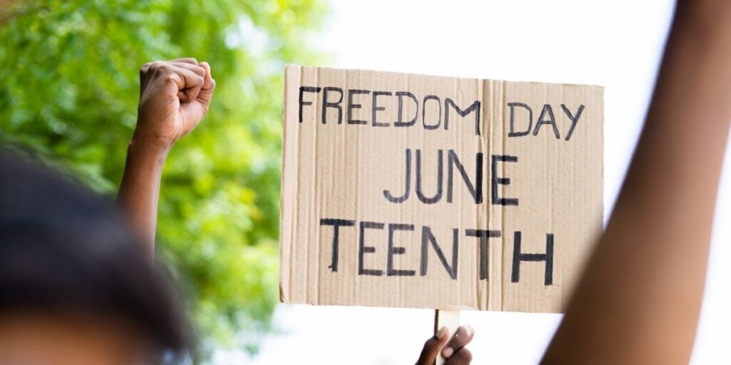 A sign reading "FREEDOM DAY JUNE TEENTH" held up outdoors, with a raised fist visible beside it.