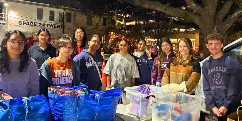 The image shows a group of nine young people standing outdoors at night, in front of a lit building. They are posing behind a table covered with large blue tote bags and transparent bins, possibly containing donations or supplies.