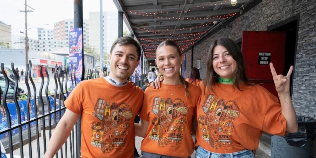 Three people in orange t-shirts with abstract designs, standing under an overhang decorated with string lights.
